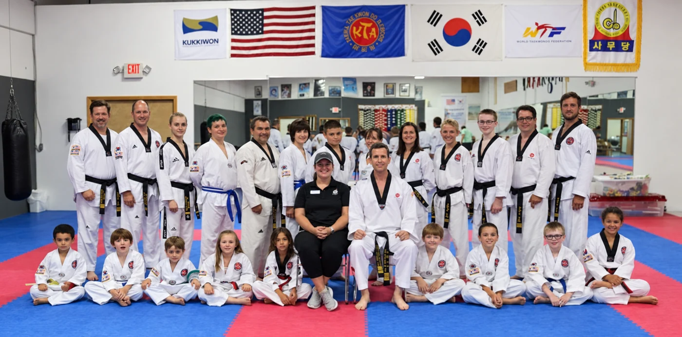 A large group of children and adults in white martial arts uniforms pose in two rows on red-and-blue mats beneath several flags in a mirrored training studio.