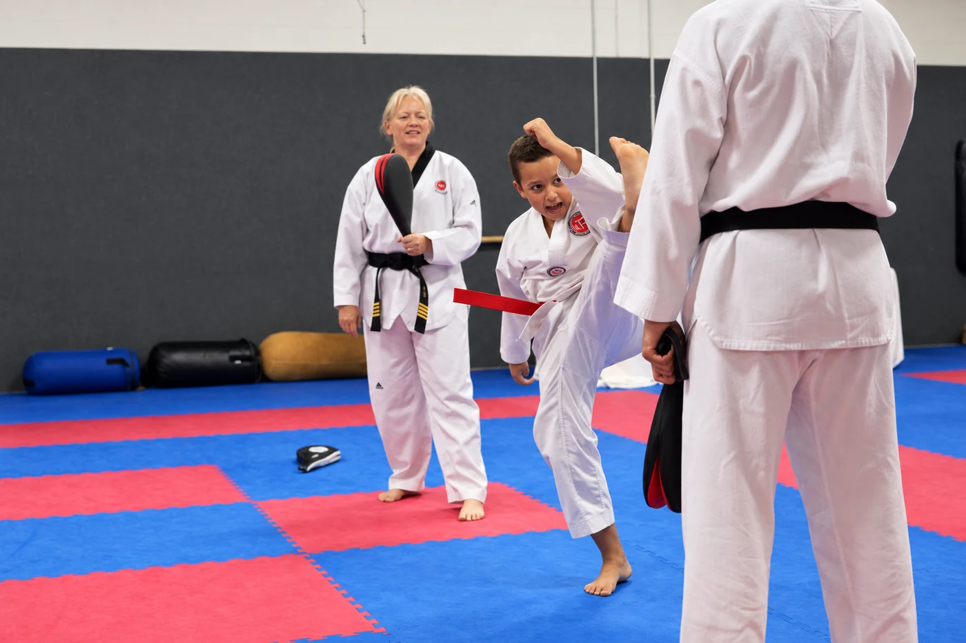 A child in a white martial arts uniform raises one leg toward a red target board while two adults and another child stand nearby on red-and-blue mats.