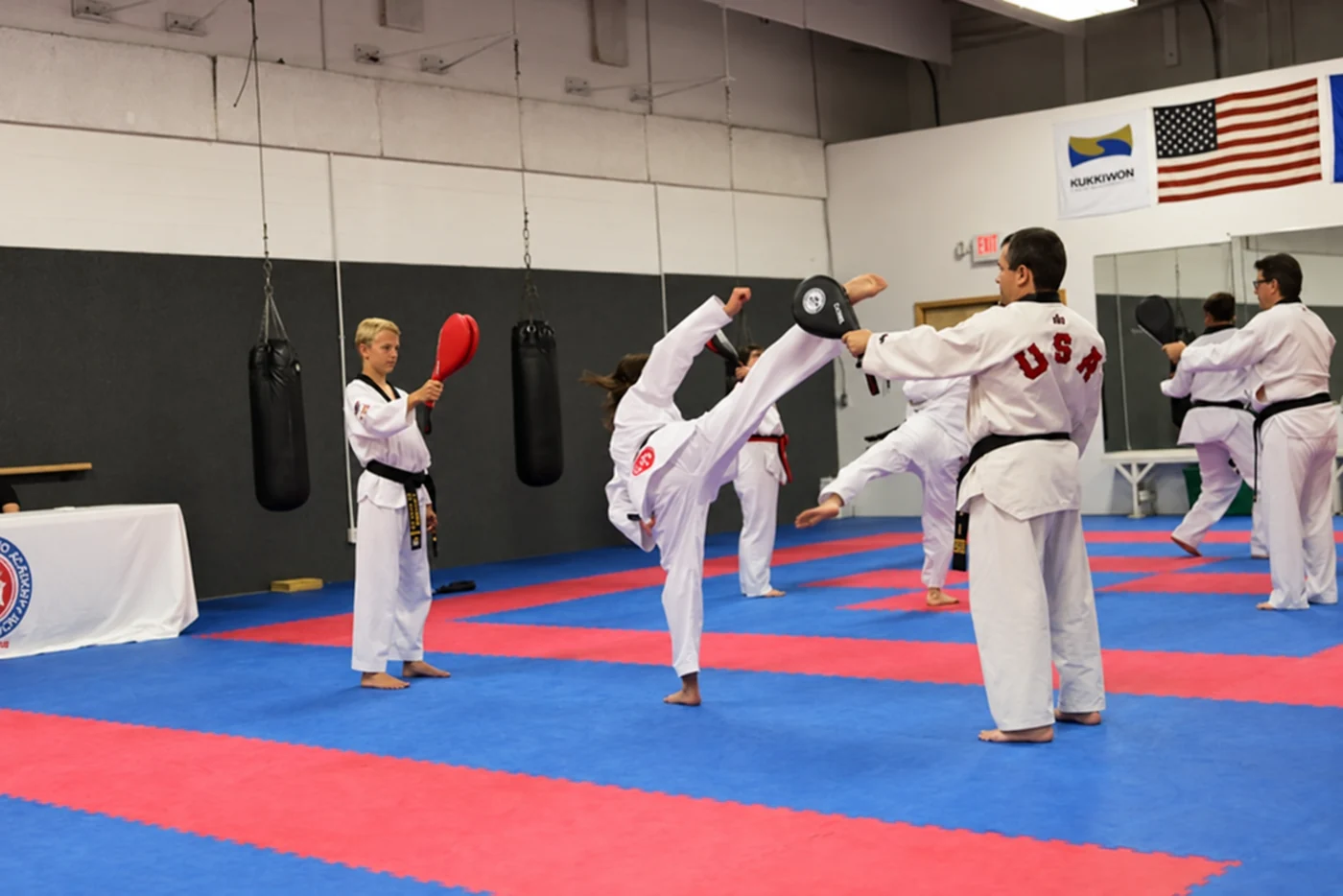 Several martial arts students practice on red-and-blue mats as one adult performs a high kick toward a handheld target and others train near mirrors and hanging punching bags.