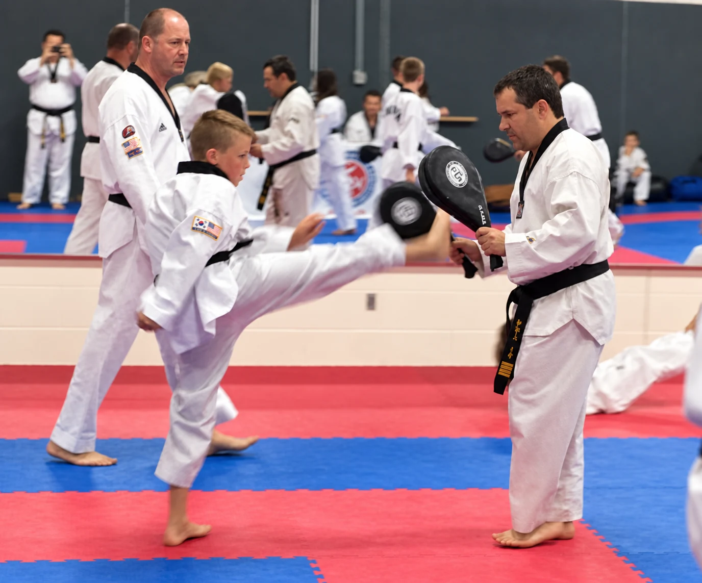 A child in a white martial-arts uniform delivers a high kick to a handheld paddle while an adult holds the target and another adult watches in a busy training hall.