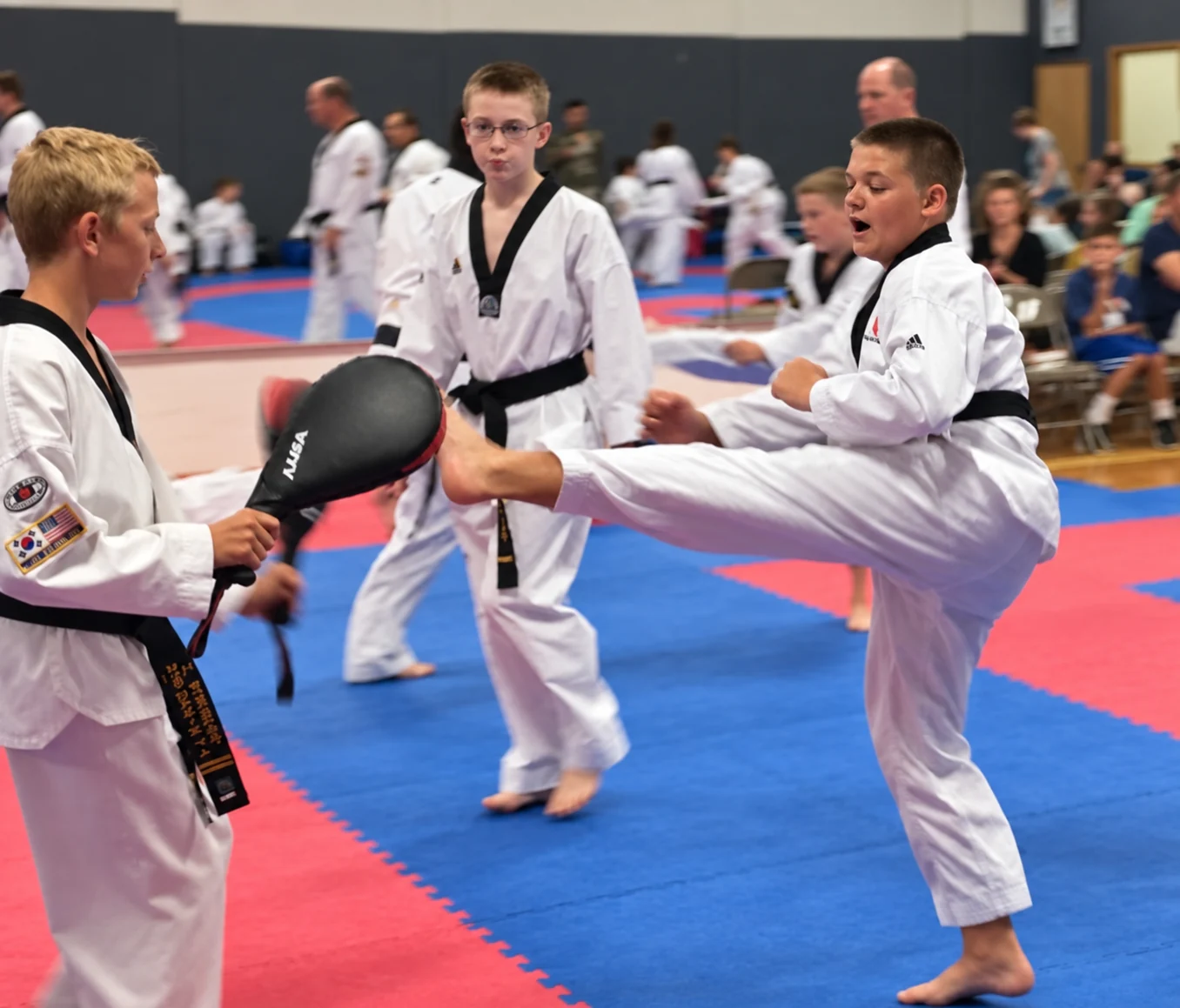 A child in a white martial-arts uniform kicks a black paddle held by another student while a child wearing glasses watches, with other participants and spectators behind them.