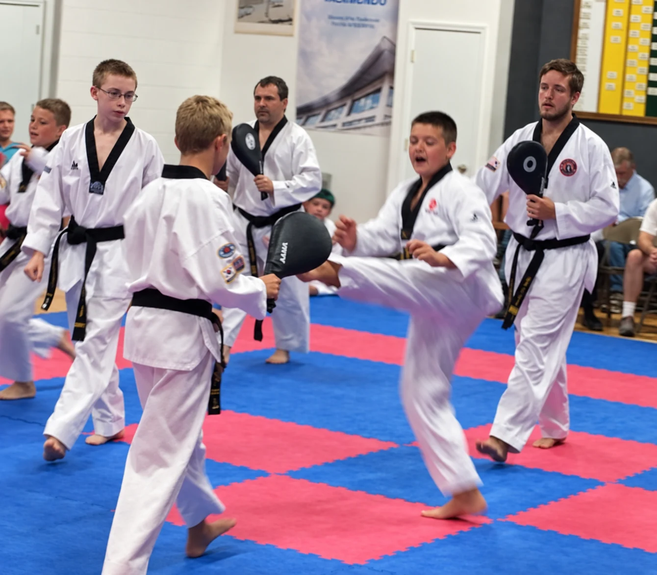 A child performs a side kick toward a handheld paddle as several children and adults in white martial-arts uniforms stand around a red-and-blue matted training area.