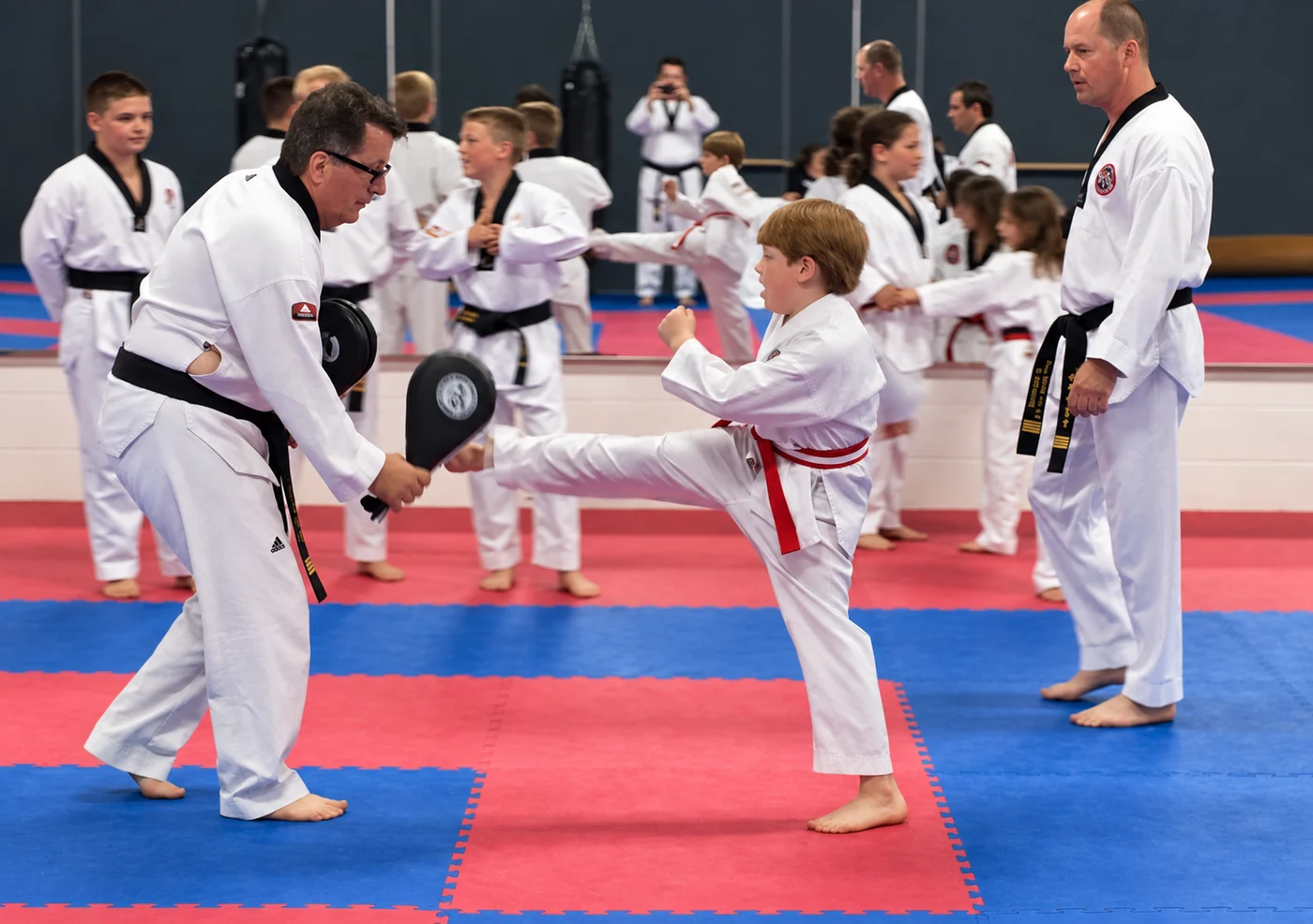 A child wearing a white martial-arts uniform and red belt kicks a black paddle held by an adult while another adult observes and a class practices in the background.