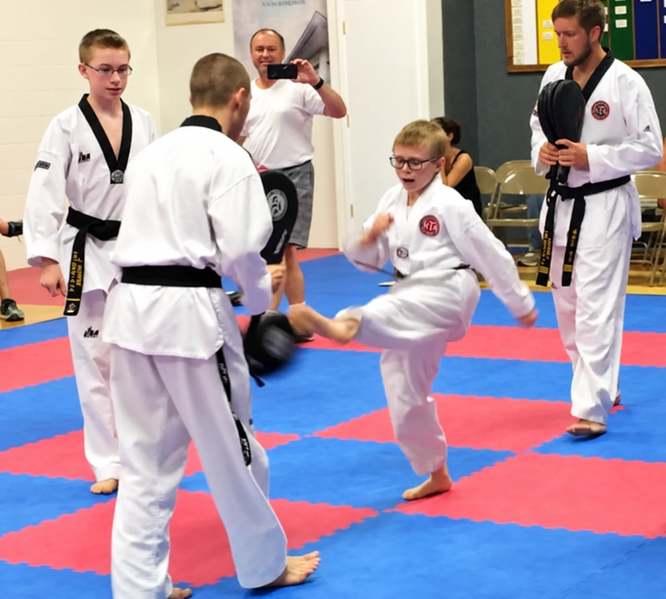 Several martial-arts students and instructors in white uniforms practice a kicking drill on red-and-blue mats while spectators watch from folding chairs.