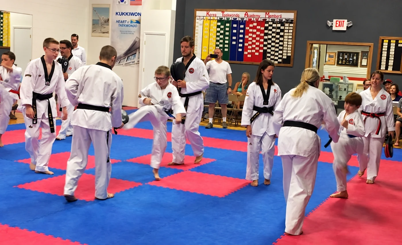 A group of children and adults in white martial-arts uniforms perform kicking drills together on red-and-blue mats inside a training hall.