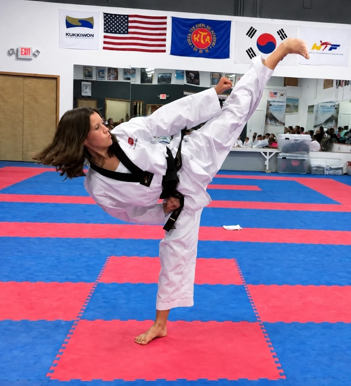 A woman in a white martial-arts uniform performs a high side kick on red-and-blue mats beneath flags and training-school banners.