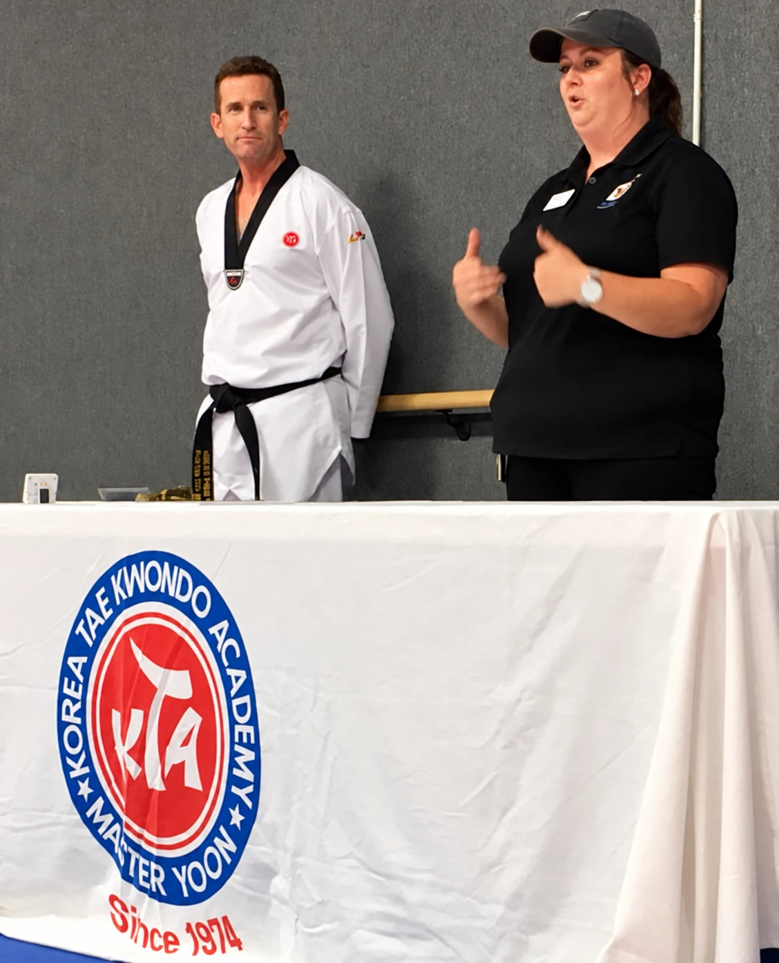 A man in a white martial-arts uniform and a woman in a black polo shirt stand behind a table covered by a cloth bearing a martial-arts academy emblem.