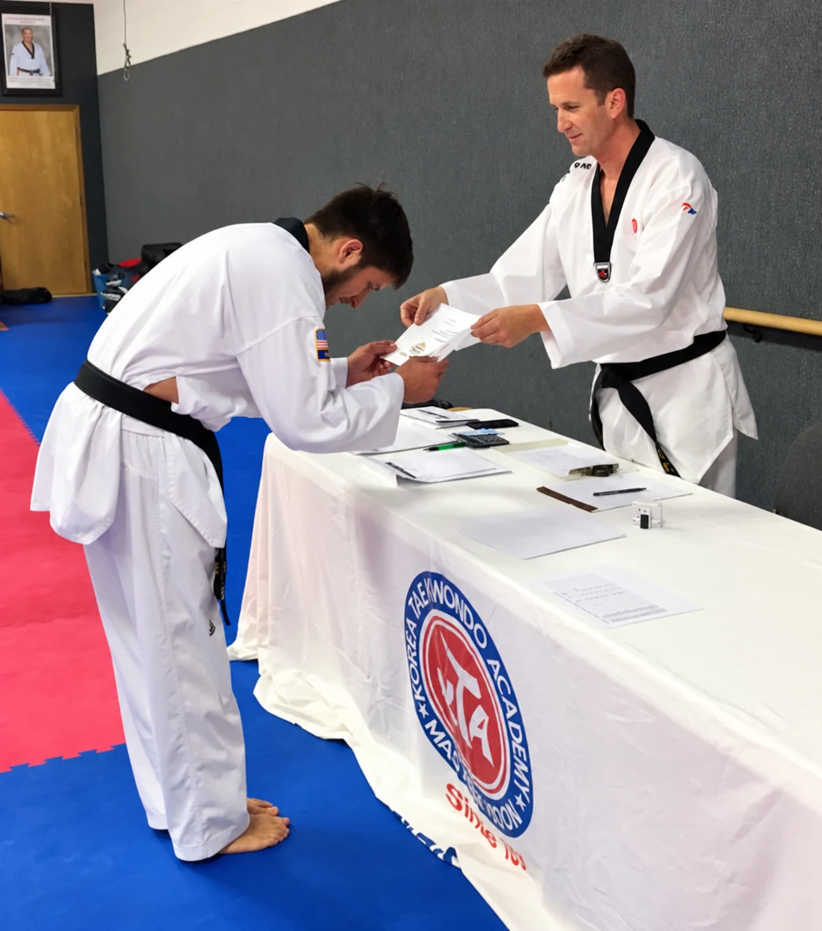 A martial arts student in a white uniform bows while receiving a certificate from another person in a white uniform and black belt behind a table covered with documents.
