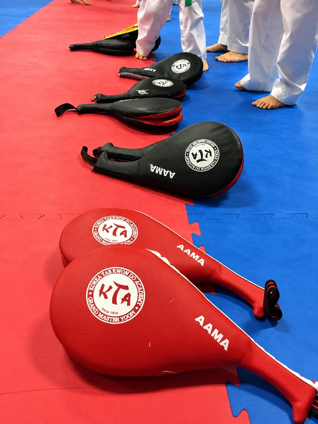 Red and black martial arts kicking paddles lie across interlocking red-and-blue floor mats near the bare feet of people wearing white uniforms.