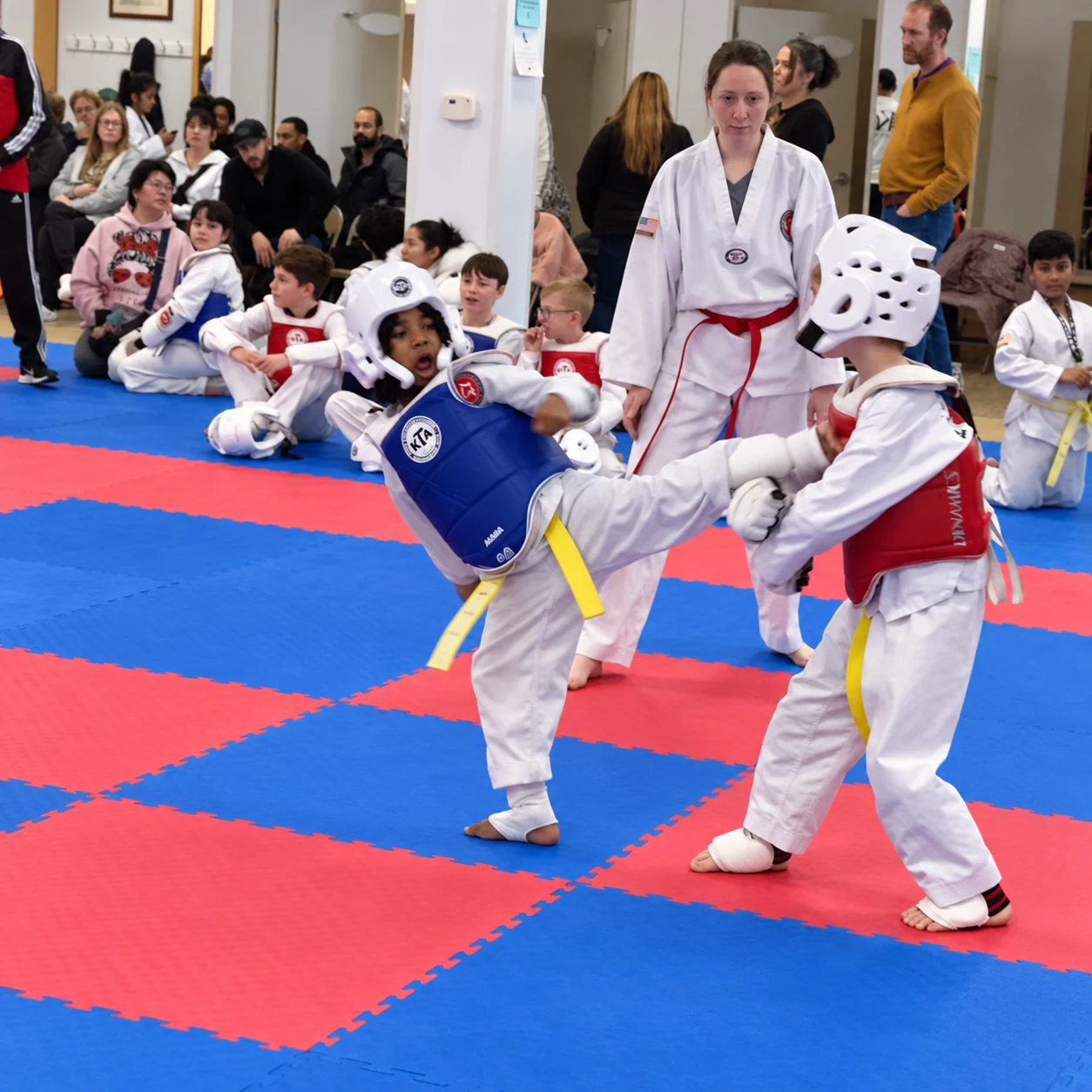 Two children wearing helmets and colored chest protectors spar on red-and-blue mats while an adult in a white uniform watches and spectators sit behind them.