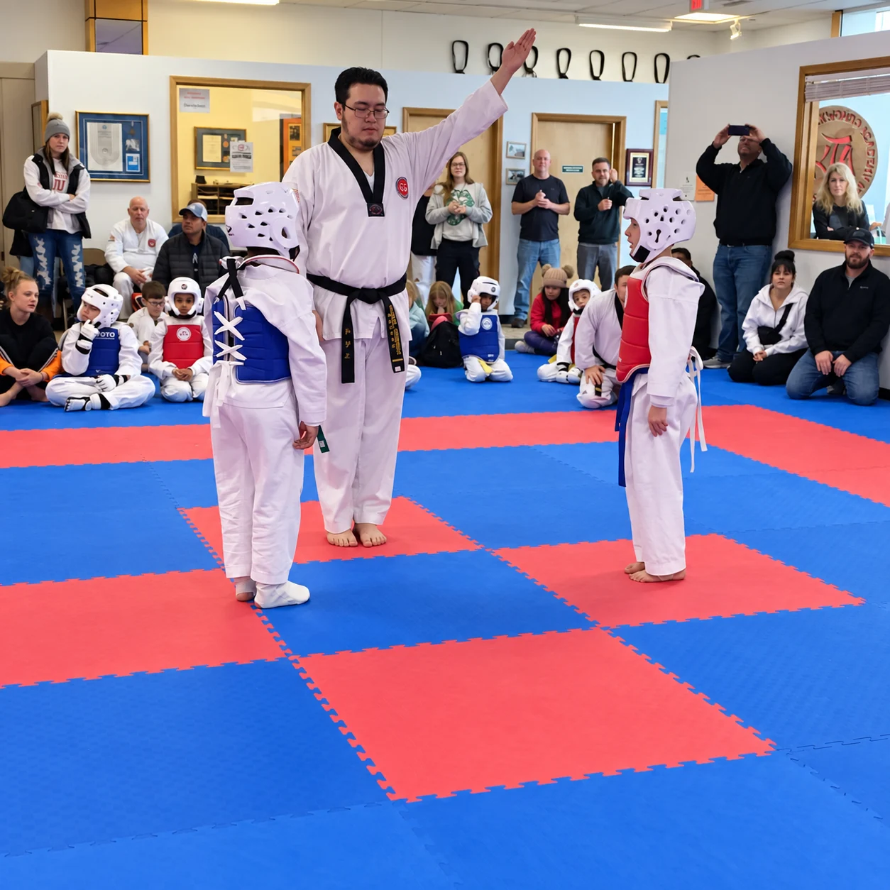 An adult in a white martial-arts uniform raises one arm while standing between two helmeted children in blue and red chest protectors, with spectators seated around the mat.