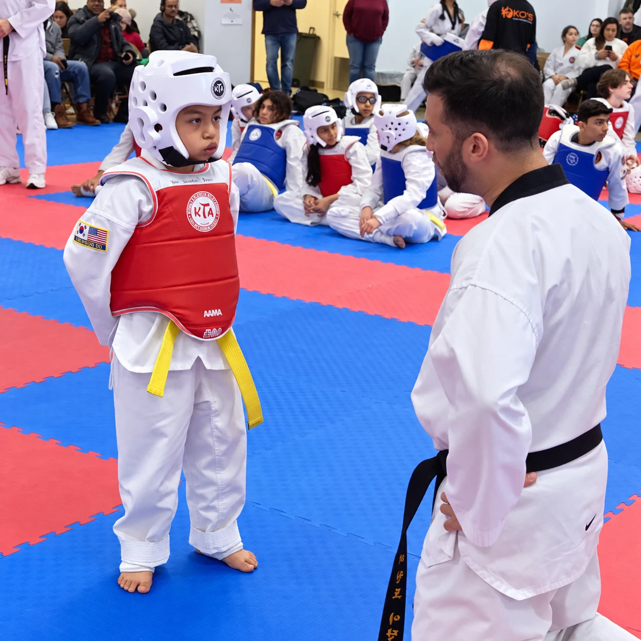 A young taekwondo student in white protective headgear, a red chest guard, and a yellow belt stands facing an instructor while other students and spectators sit behind them.