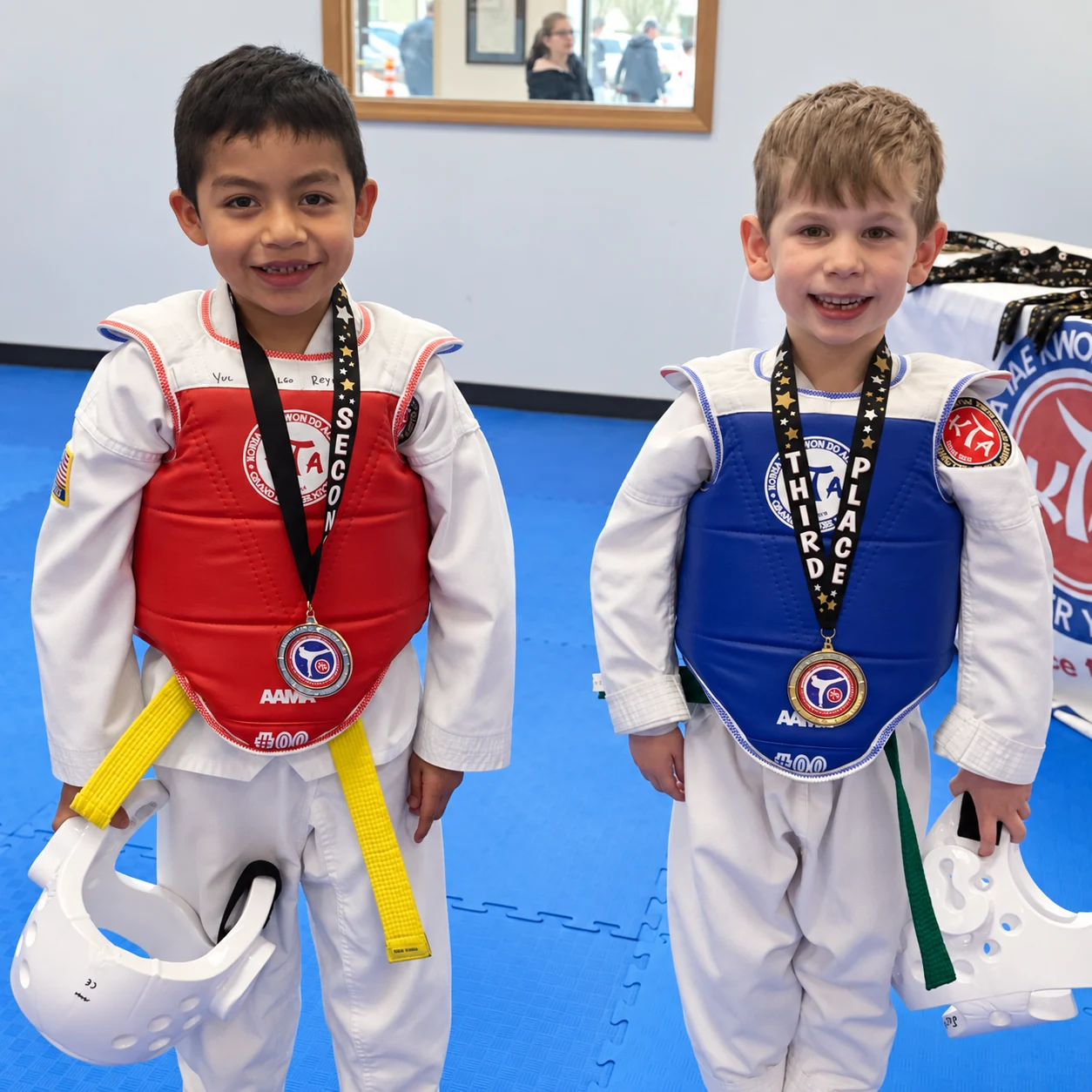 Two smiling children in white taekwondo uniforms and red and blue chest guards stand on a blue mat wearing medals and holding white protective helmets.