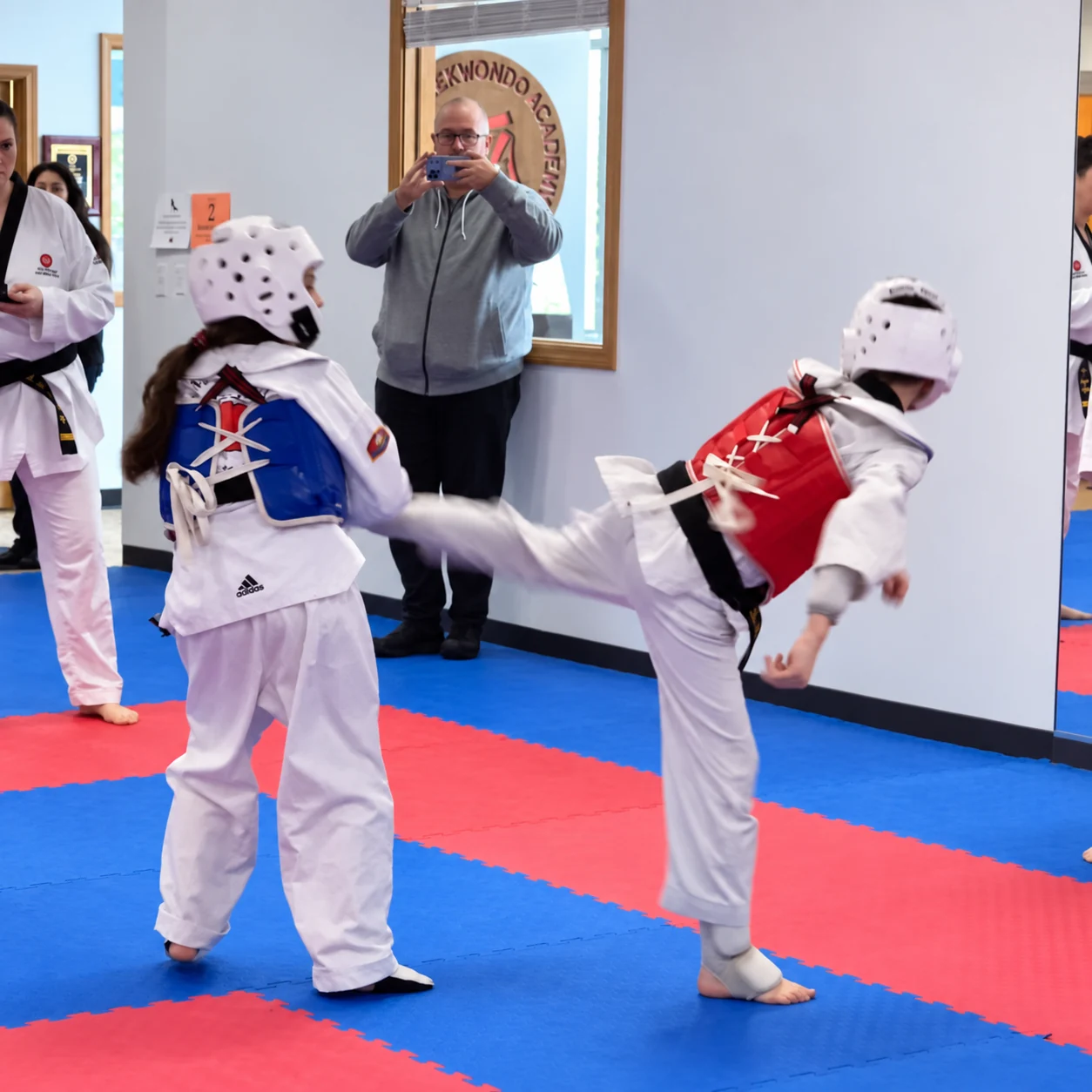 Two young taekwondo competitors in white uniforms and protective helmets spar on red-and-blue mats as one delivers a side kick toward the other’s blue chest guard.
