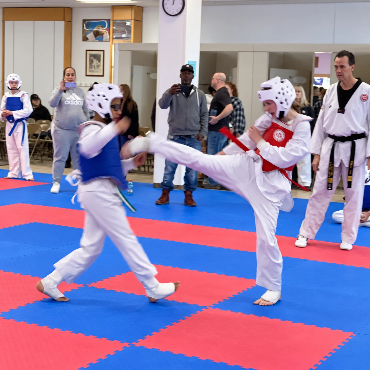 Two young taekwondo competitors spar on red-and-blue mats, with one in a red chest guard extending a high kick toward an opponent in a blue chest guard while adults watch nearby.