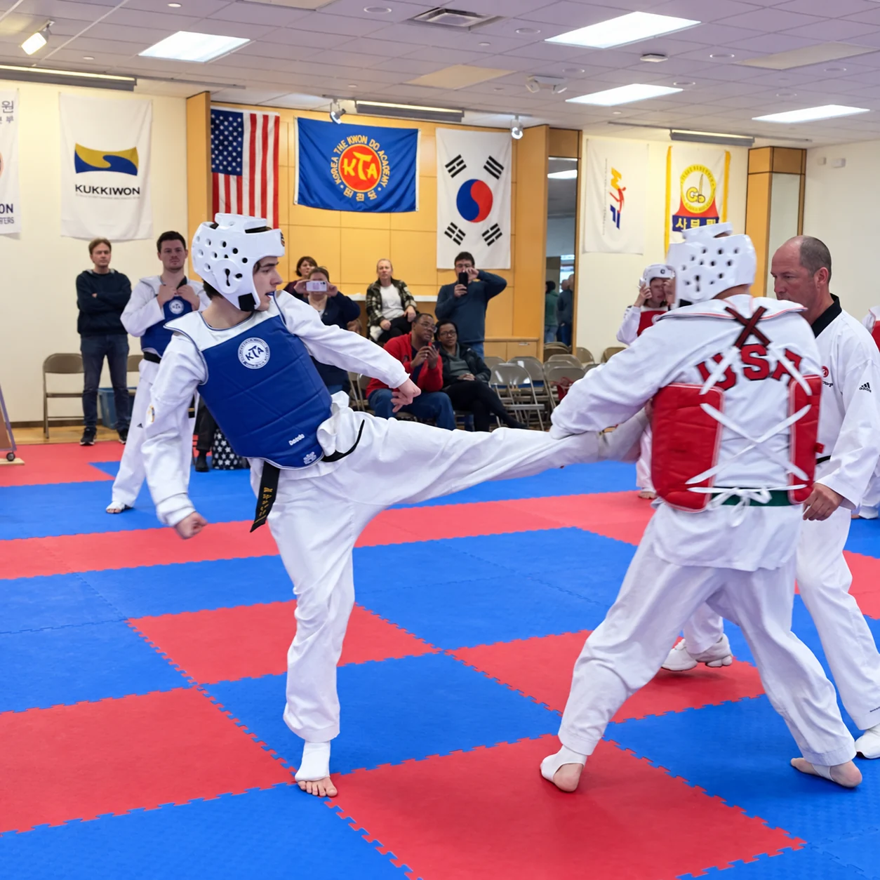Two martial artists wearing white uniforms, head guards, and blue and red chest protectors spar on red-and-blue mats while an official and spectators watch inside a training hall.