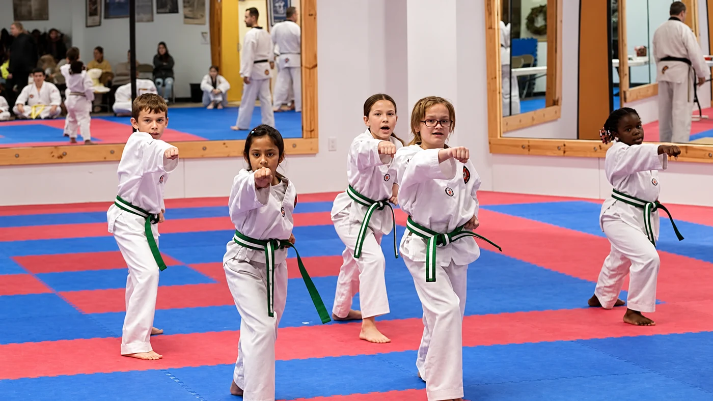 Five children in white martial-arts uniforms practice synchronized punches on red-and-blue mats in a mirrored training room.
