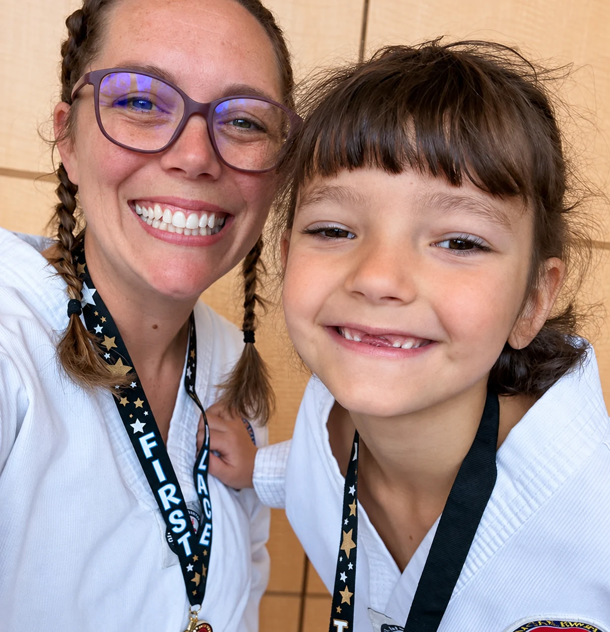 Close-up selfie of a smiling adult and child wearing white martial-arts uniforms and medal lanyards indoors.