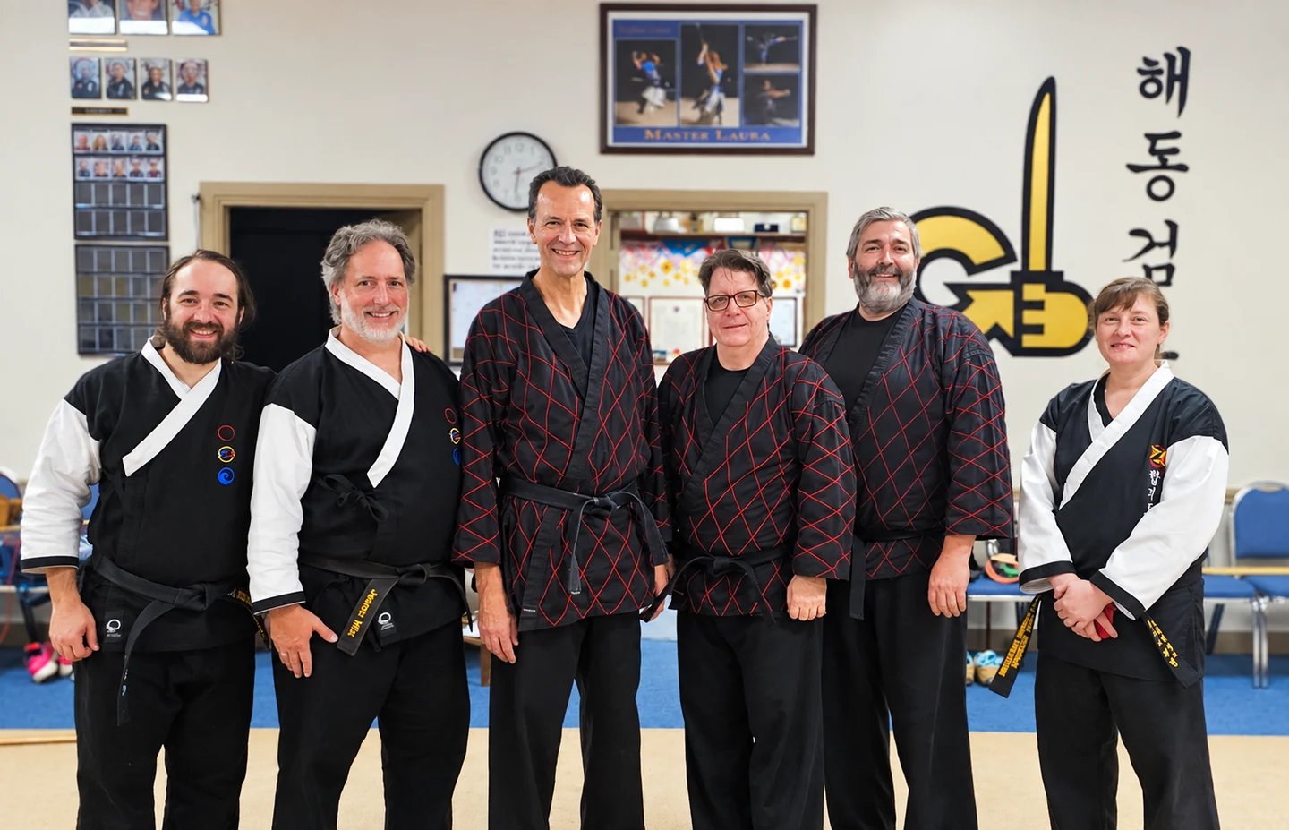 Six martial arts practitioners in black uniforms posing together inside a training studio.